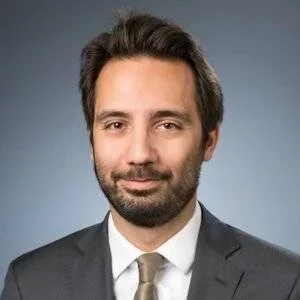Man with short dark hair and a trimmed beard wearing a suit and tie against a neutral background.