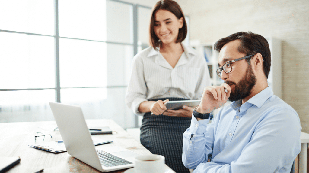 busines professionals in office looking at laptop on desk, woman holding tablet smiling, man looking thoughtful seated at desk