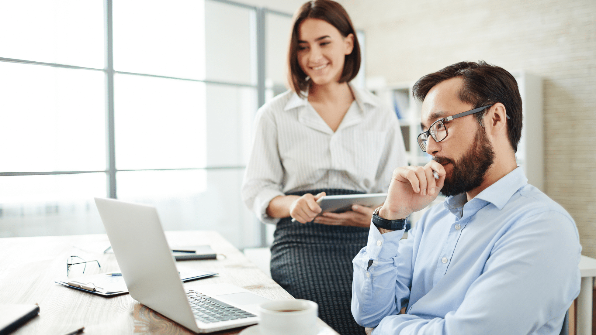 busines professionals in office looking at laptop on desk, woman holding tablet smiling, man looking thoughtful seated at desk