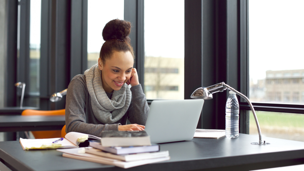 Female college student in library study area on laptop with books smiling and reading the news